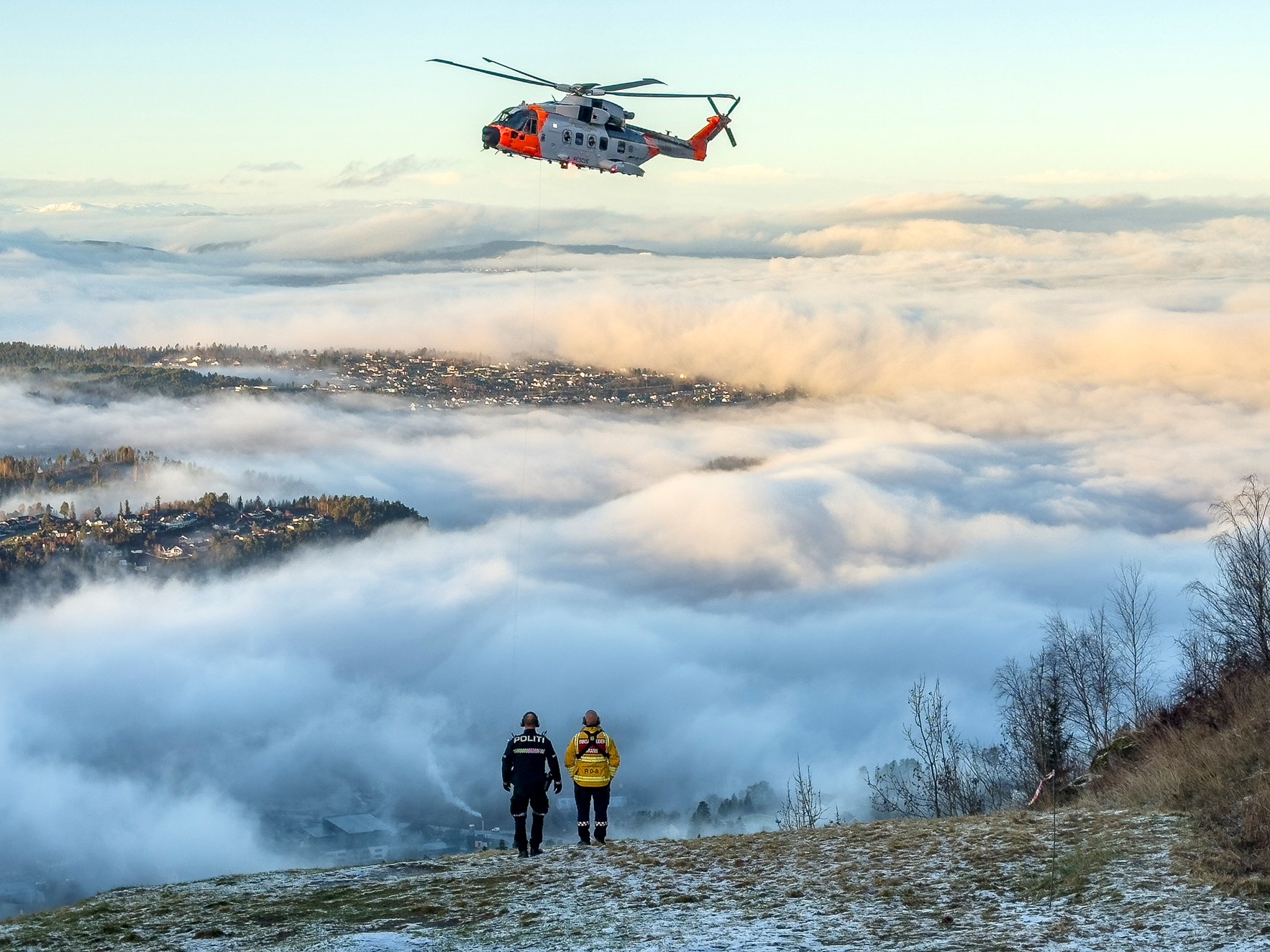 Et redningshelikopter henger lavt over en bratt skr&aring;ning med utsikt over hus som er delvis skjult i skodde. En politimann og en brannmann st&aring;r i forgrunnen.