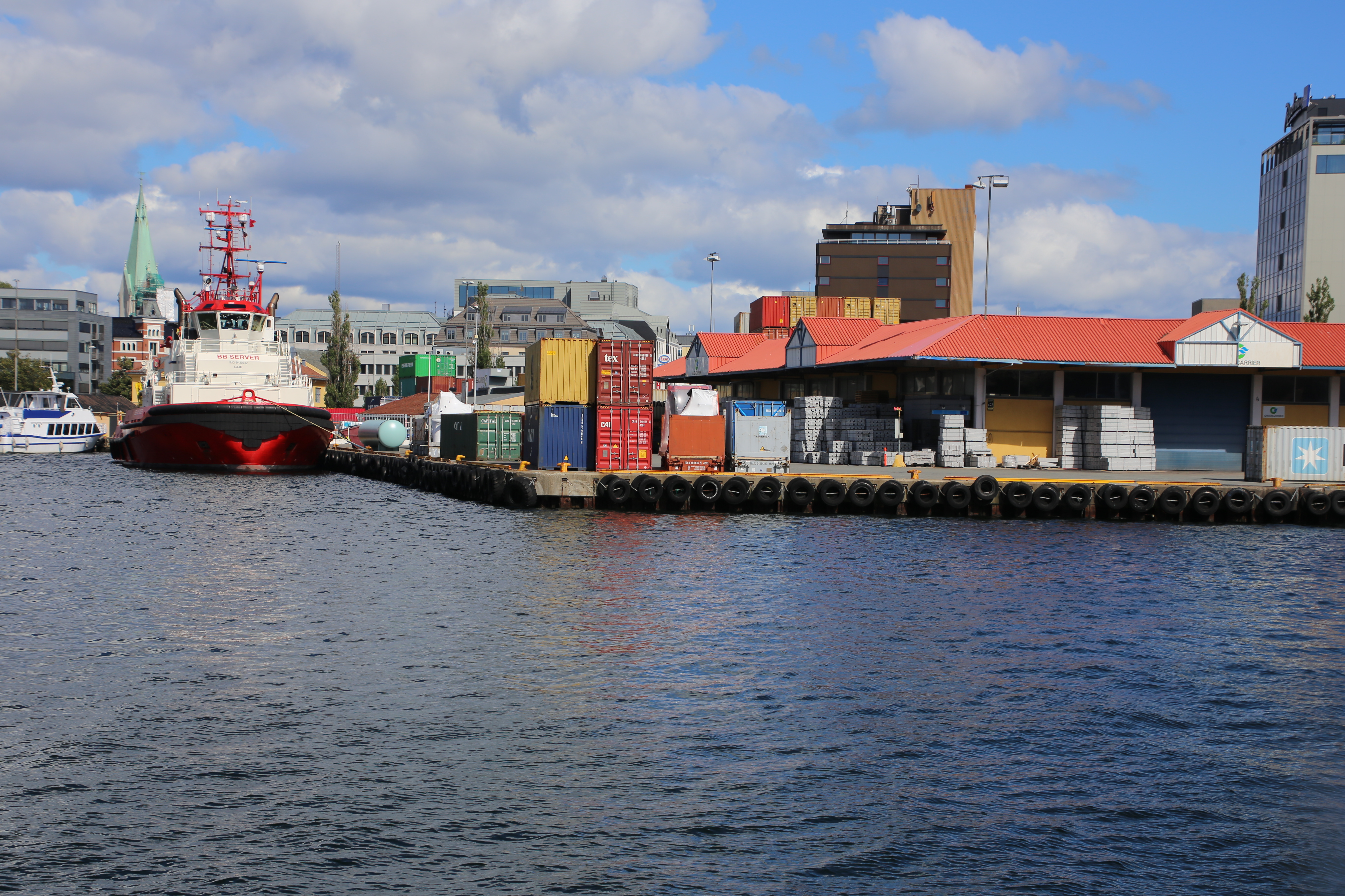Kaianlegg i Kristiansand havn med konteinere og paller med varer på kaikanten, båter til venstre
