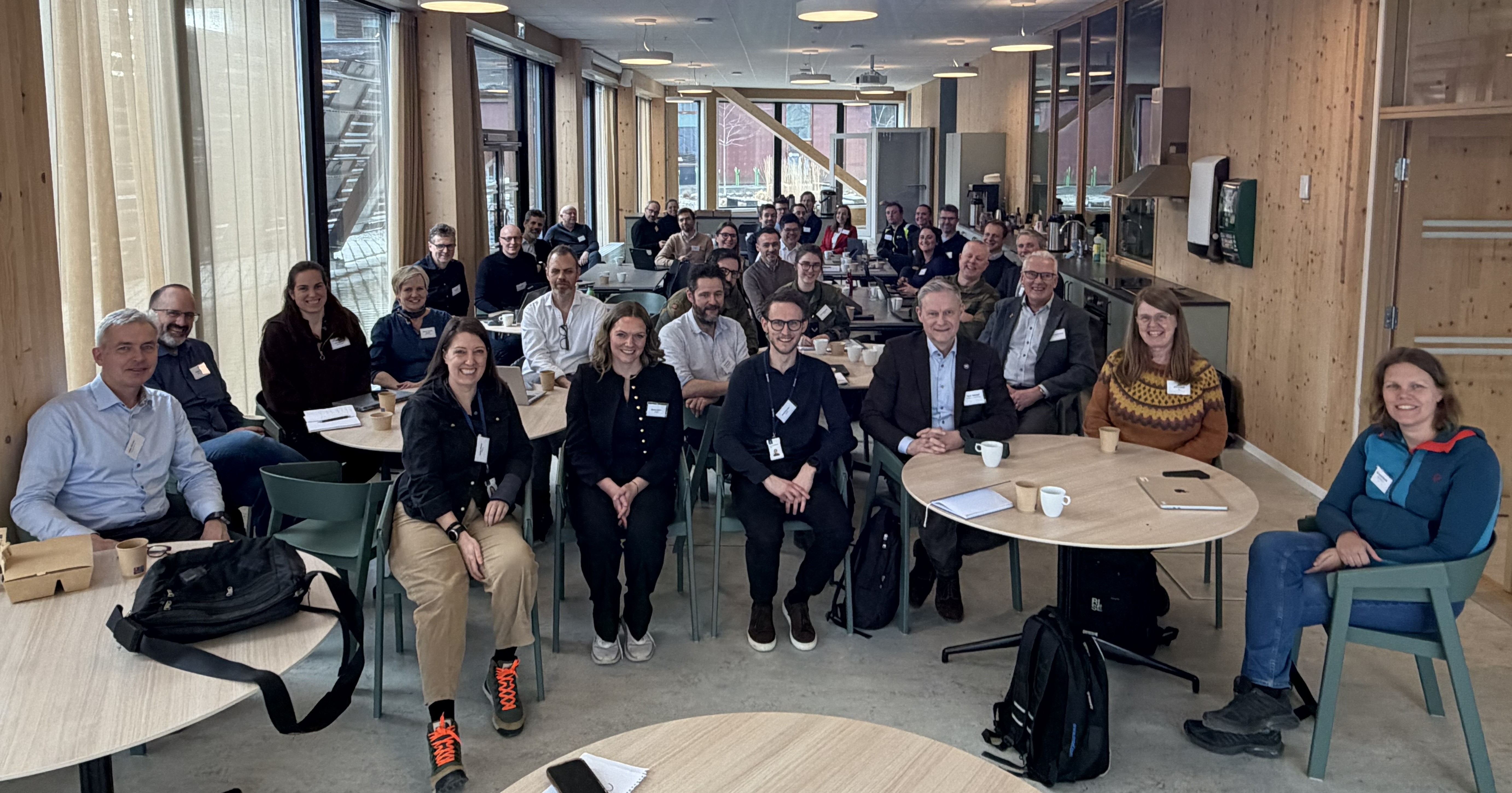 Fellesbilde med smilende mennesker med navneskilt sittende ved runde bord i m&oslash;te.