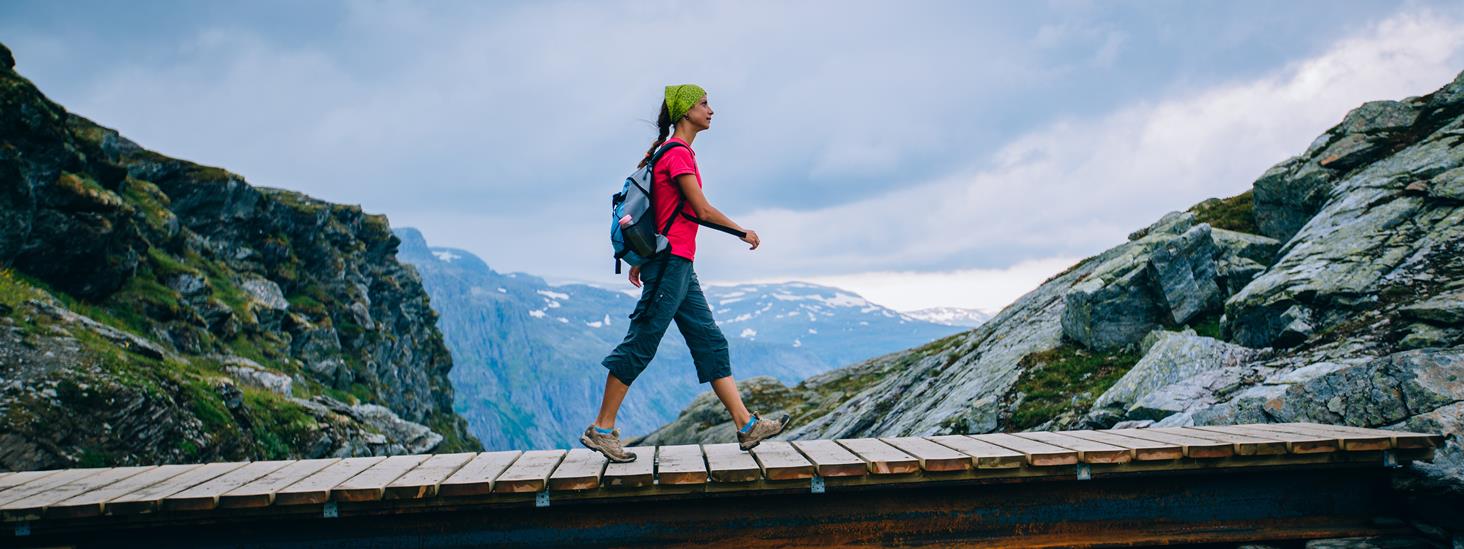 Ung sporty kvinne vandrer på steinete sti som krysser bekk/elv på trebro
