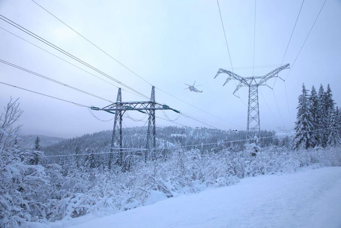 H&oslash;yspentmast og ledningsnett med et helikopter svevende over (foto Wilhelm B&oslash;e/ Kartverket)