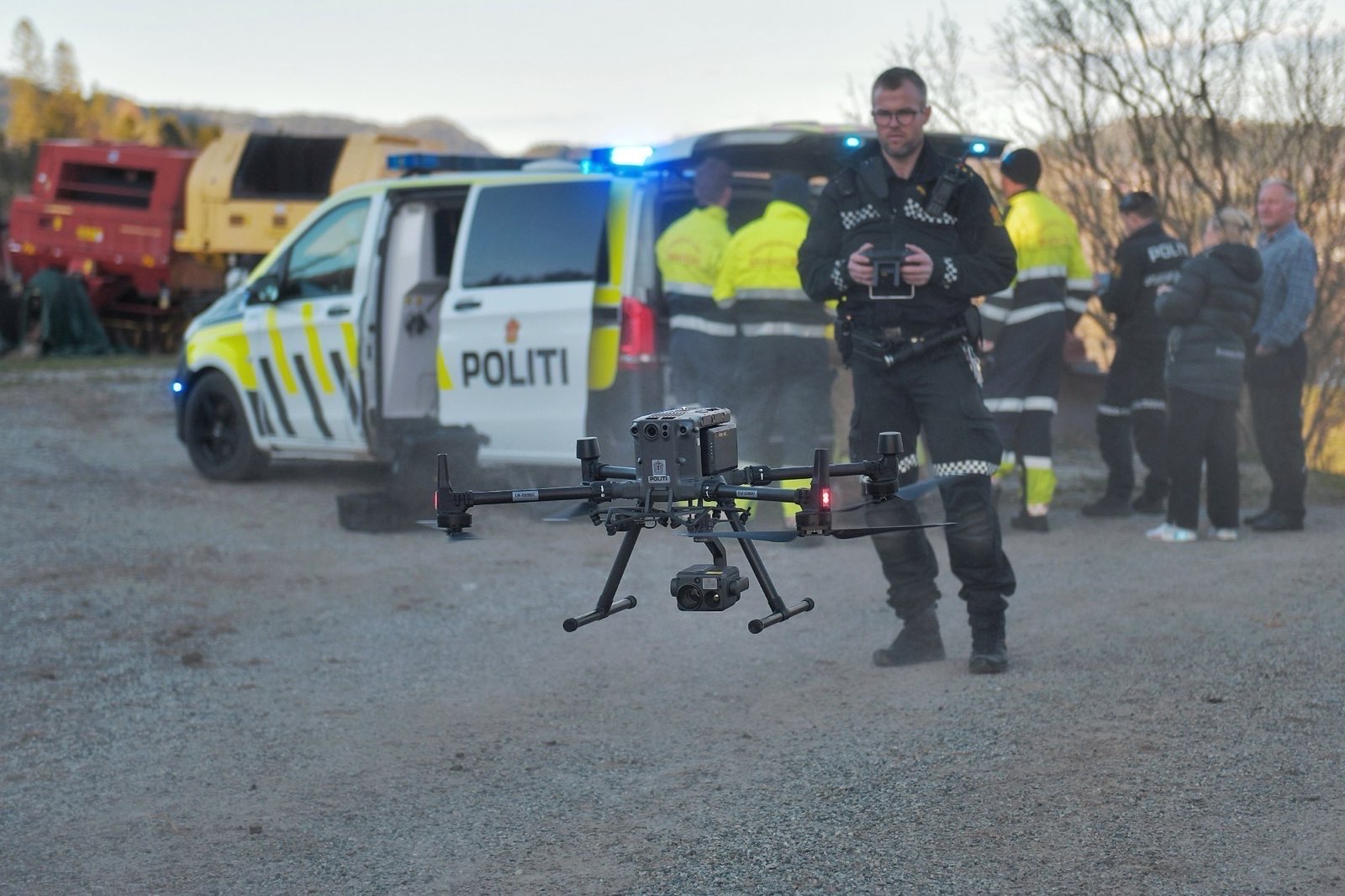 Politibetjent opererer en drone, med utrykningskj&oslash;ret&oslash;y og flere tjenestepersoner i bakgrunnen.