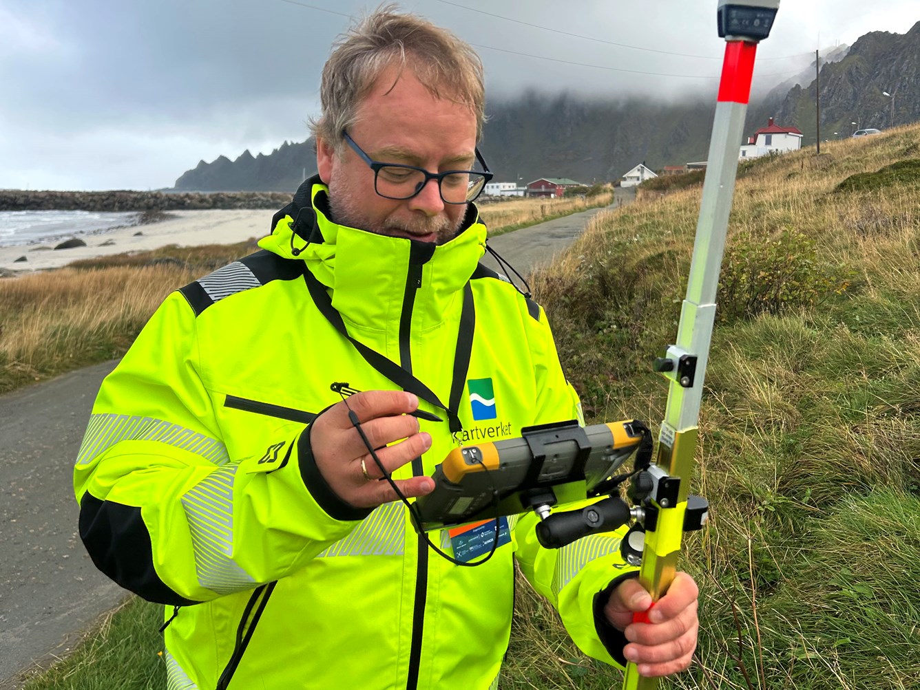 En person i gul jakke st&aring;r ute ved en GPS-stasjon st&aring;r ved en vei, med strand og fjell i bakgrunnen.