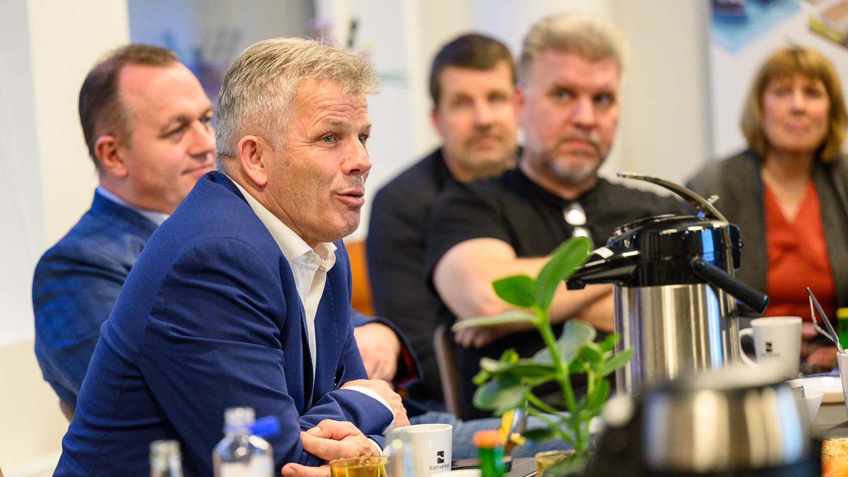 Fem mennesker sitter ved et bord dekt med, blomst, glass, krus, frukt og temokanner.