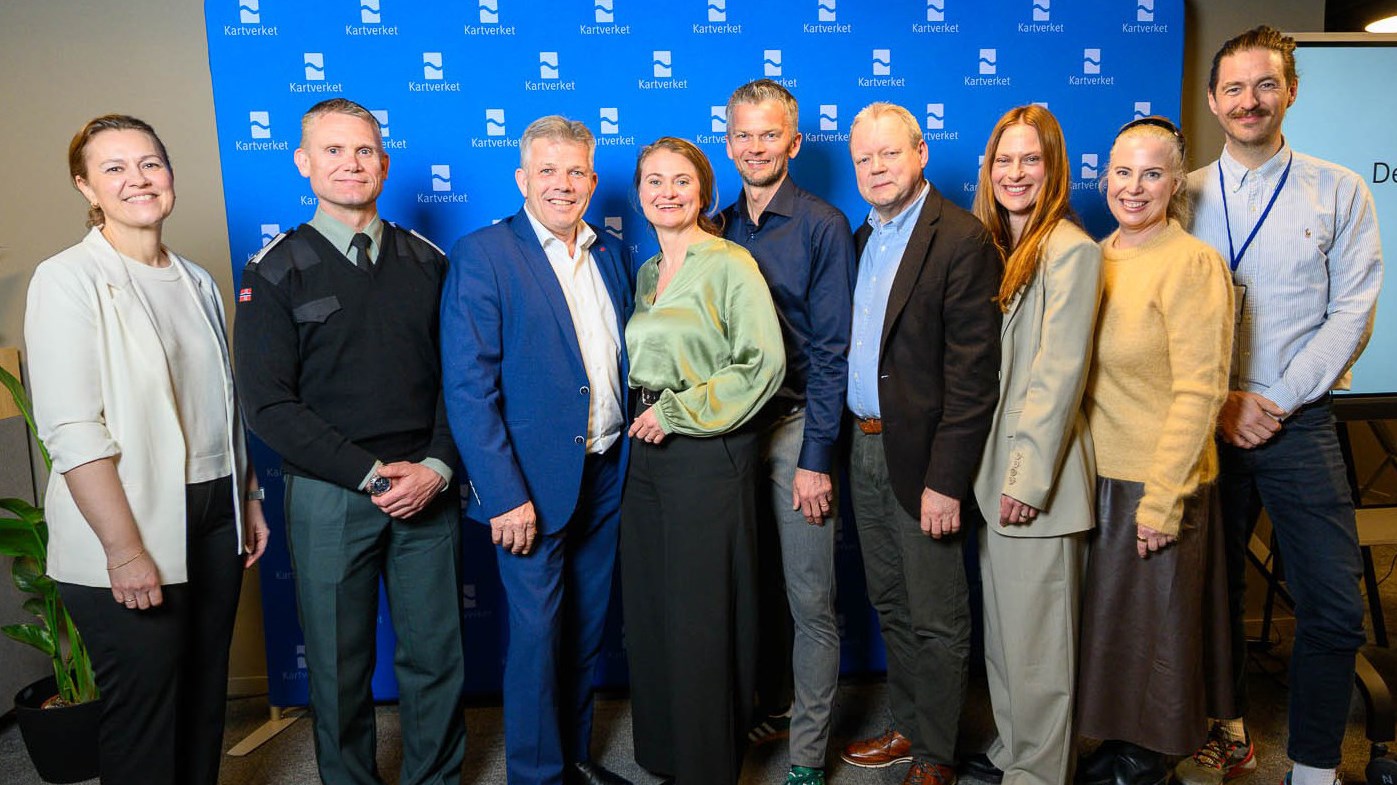 Ni glade mennesker st&aring;r oppstilt foran en bl&aring; fotovegg med Kartverkets logo.