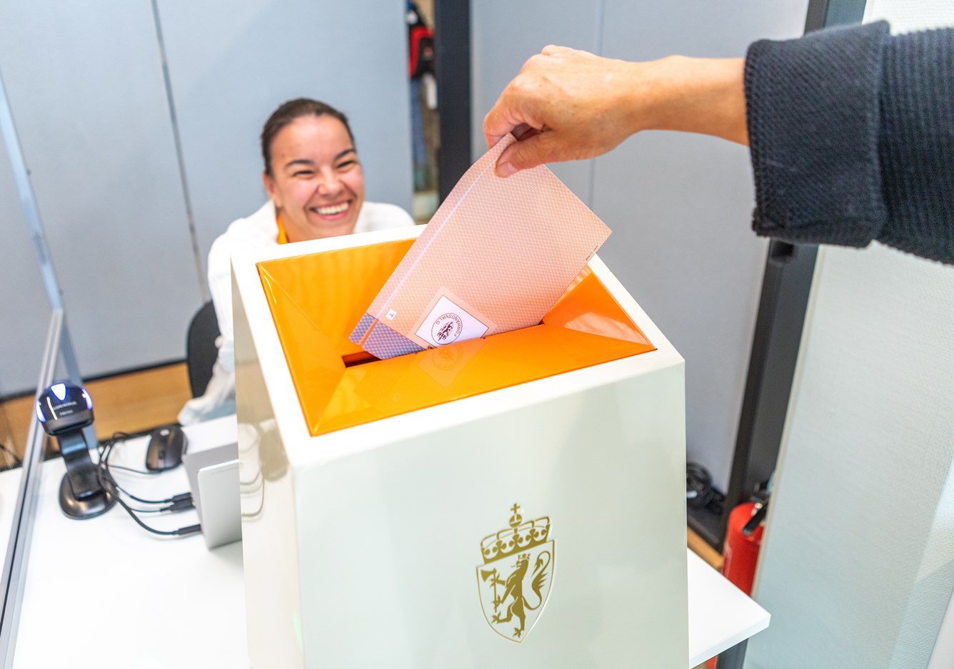 En stemmeseddel legges oppi ei valgurne. Bak valgurna sitter ei blid dame.