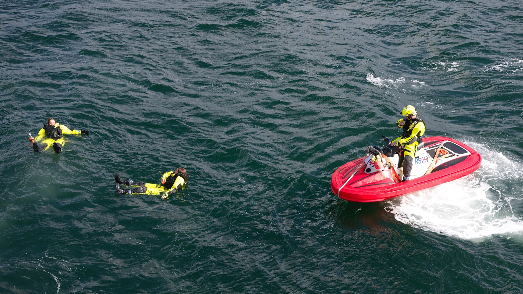 To mark&oslash;rer i flyedrakt ligger i vannet mens en vannscooter fra Redningsselskapet er p&aring; vei bort for &aring; redde dem.
