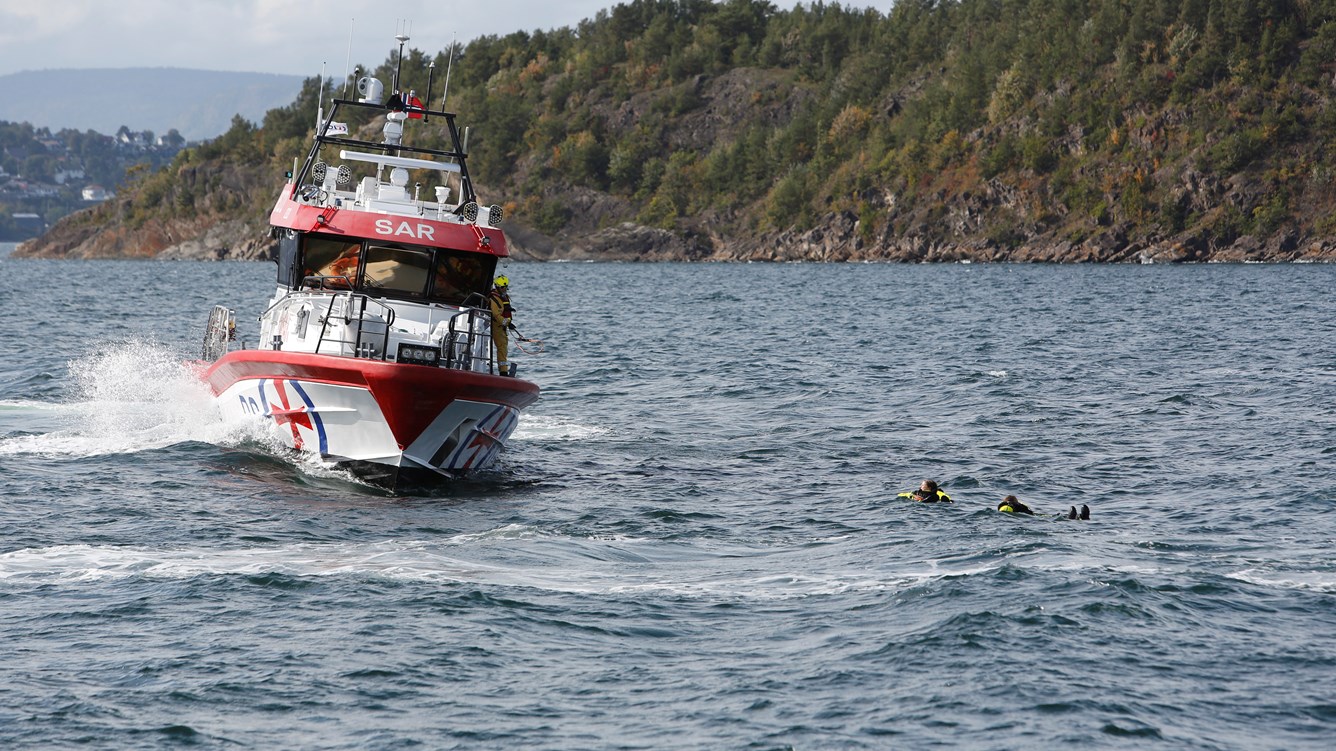 Redningssk&oslash;yte p&aring; vei for &aring; redde to personer i flytedrakt som ligger i vannet.