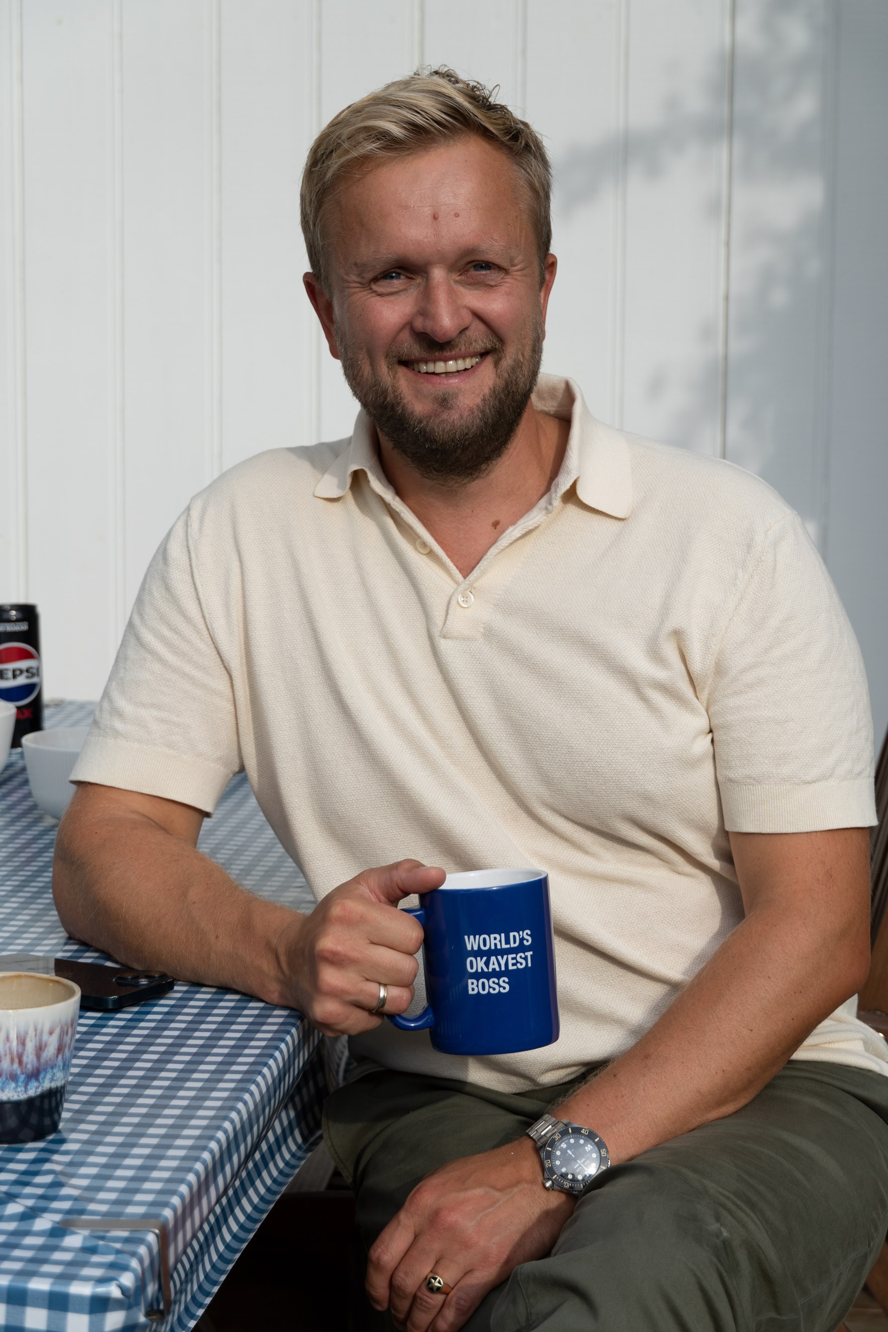 Mann med blondt h&aring;r og skjegg holder en bl&aring; kopp mens han smiler inn i kamera