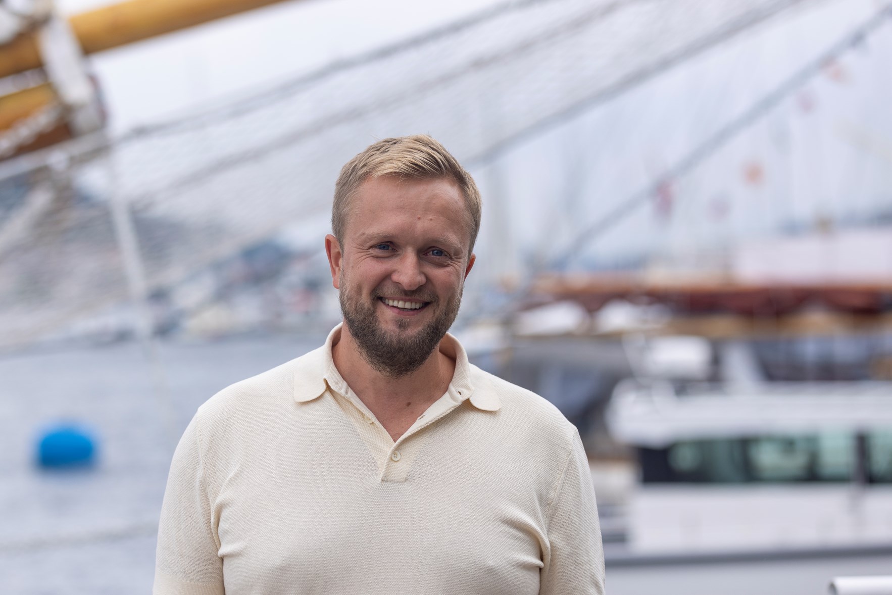 Mann med blondt h&aring;r, m&oslash;rkt skjegg og hvit skjorte smiler inn i kamera.