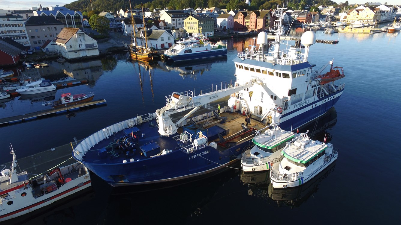 Luftfoto av havn med et stort bl&aring;tt forskningsskip fort&oslash;yd ved kai, omgitt av mindre b&aring;ter. P&aring; land ligger tettbygde hus i ulike farger, og vannet er rolig med speilinger av bygninger og fart&oslash;y.