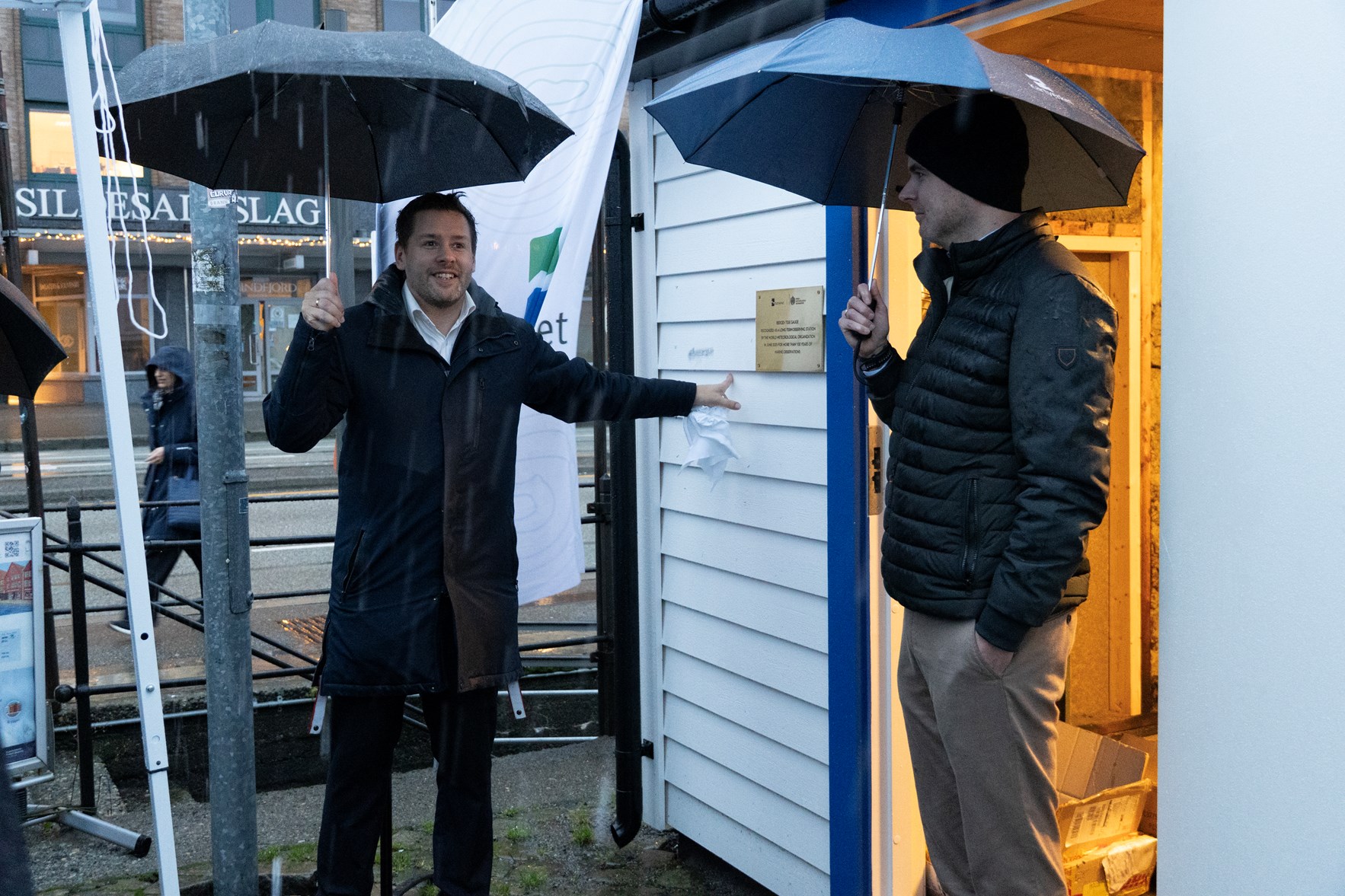 To menn st&aring;r foran et lite hvitt bygg p&aring; ei kai i Bergen. Begge holder en paraply.