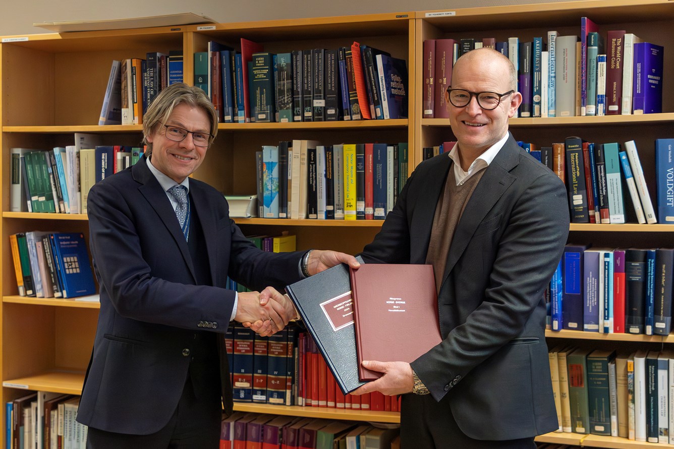 To menn foran en bokhylle tar hverandre i hendene mens de smiler til kamera. En av mennene holder to b&oslash;ker i sin venstre h&aring;nd. 